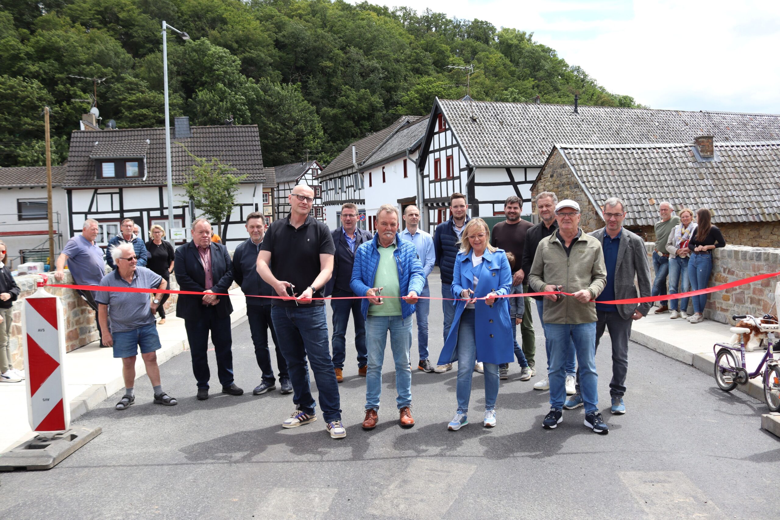 Mehrere Menschen zerschneiden ein Band, um die Brücke in Iversheim einzuweihen. In im Hintergrund mehr Menschen und Fachwerkhäuser.